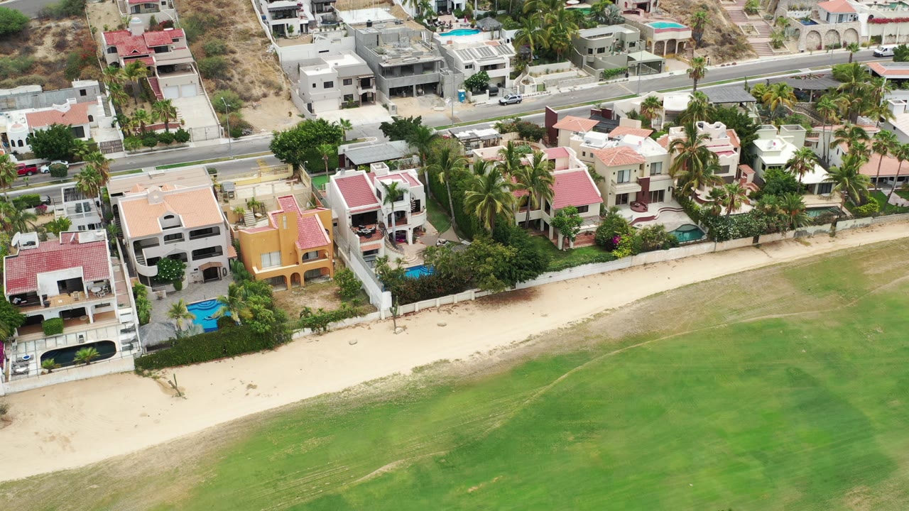 Los Cabos aerial frame centered on a row of coastal homes, palm trees, small pools, pale sand, green water, and a paved road behind the properties with no visible people.
