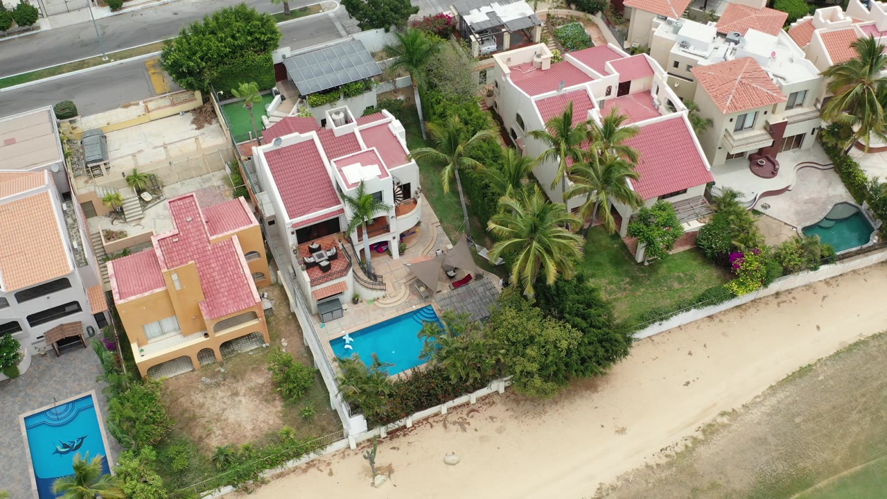 Los Cabos residential aerial with a central pink-roofed house, bright blue pools at the lower edges, palms, garden walls, and no visible people.