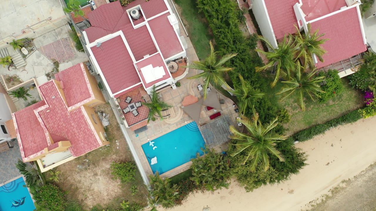 Overhead view of red-roofed homes, blue pool, palms, and sandy ground.
