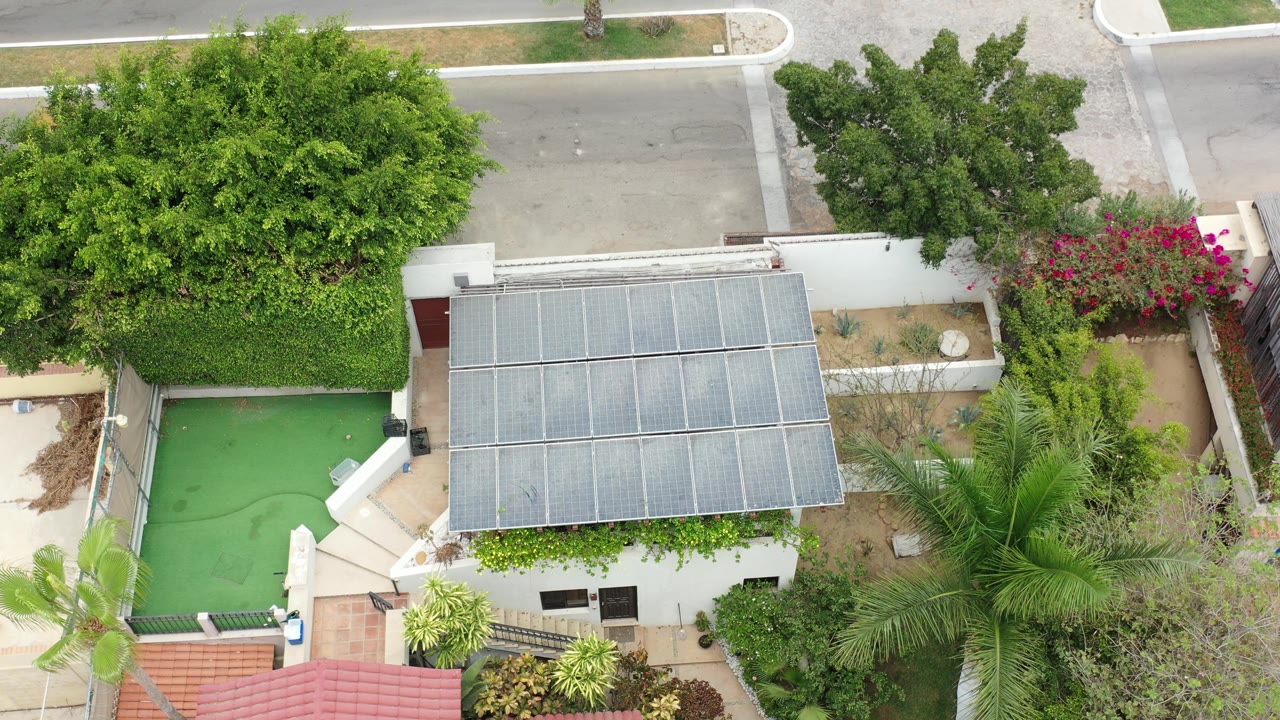 Overhead frame in Los Cabos, Mexico showing a white residential building with a gray panel-covered roof, green courtyard surface, hedges, palms, flowering shrubs, and no visible people.