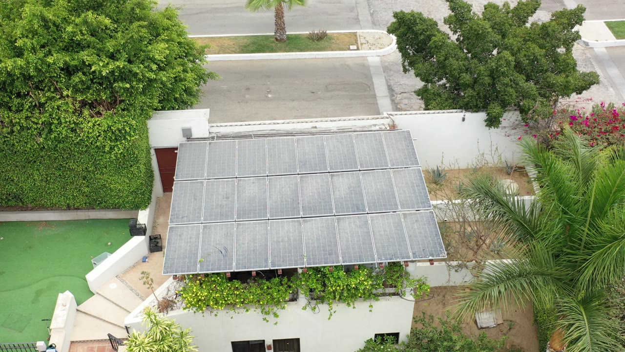 Aerial view of a white building entrance with a metal canopy, hedges, palm fronds, and a road above.