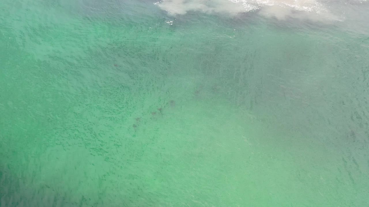 A high overhead Los Cabos water frame shows pale green-blue shallows, scattered darker areas, and foamy white wave wash at the top with no visible people.