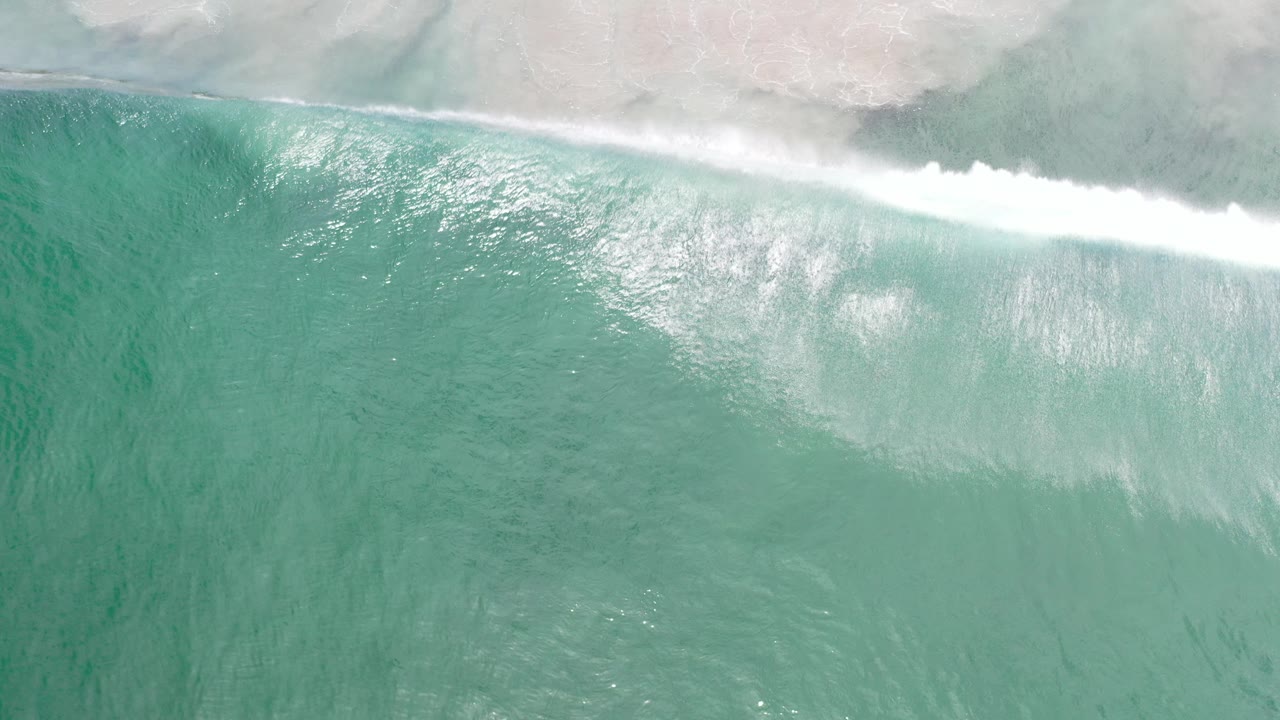 Overhead view of turquoise ocean in Los Cabos, Mexico with a white breaking wave along the upper edge and no visible people.