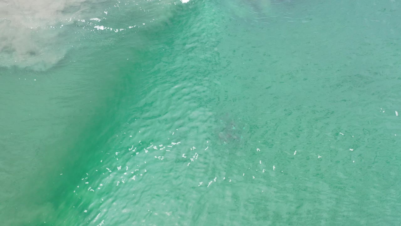 Green shallow seawater in Los Cabos with white surf foam entering from the upper left edge, sandy texture beneath the surface, and no visible people.