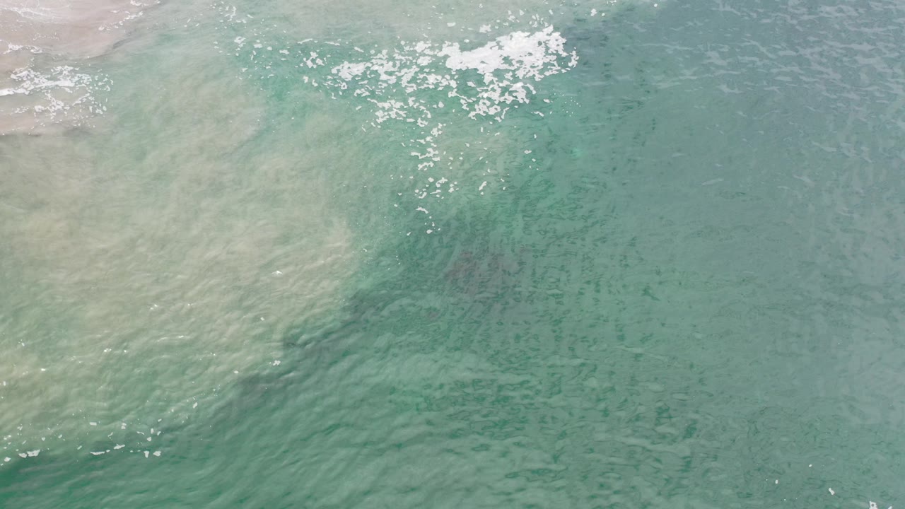 Overhead view of turquoise ocean water in Los Cabos, Mexico, with pale shallow water at the upper left, scattered white seafoam near the top, and no visible people.