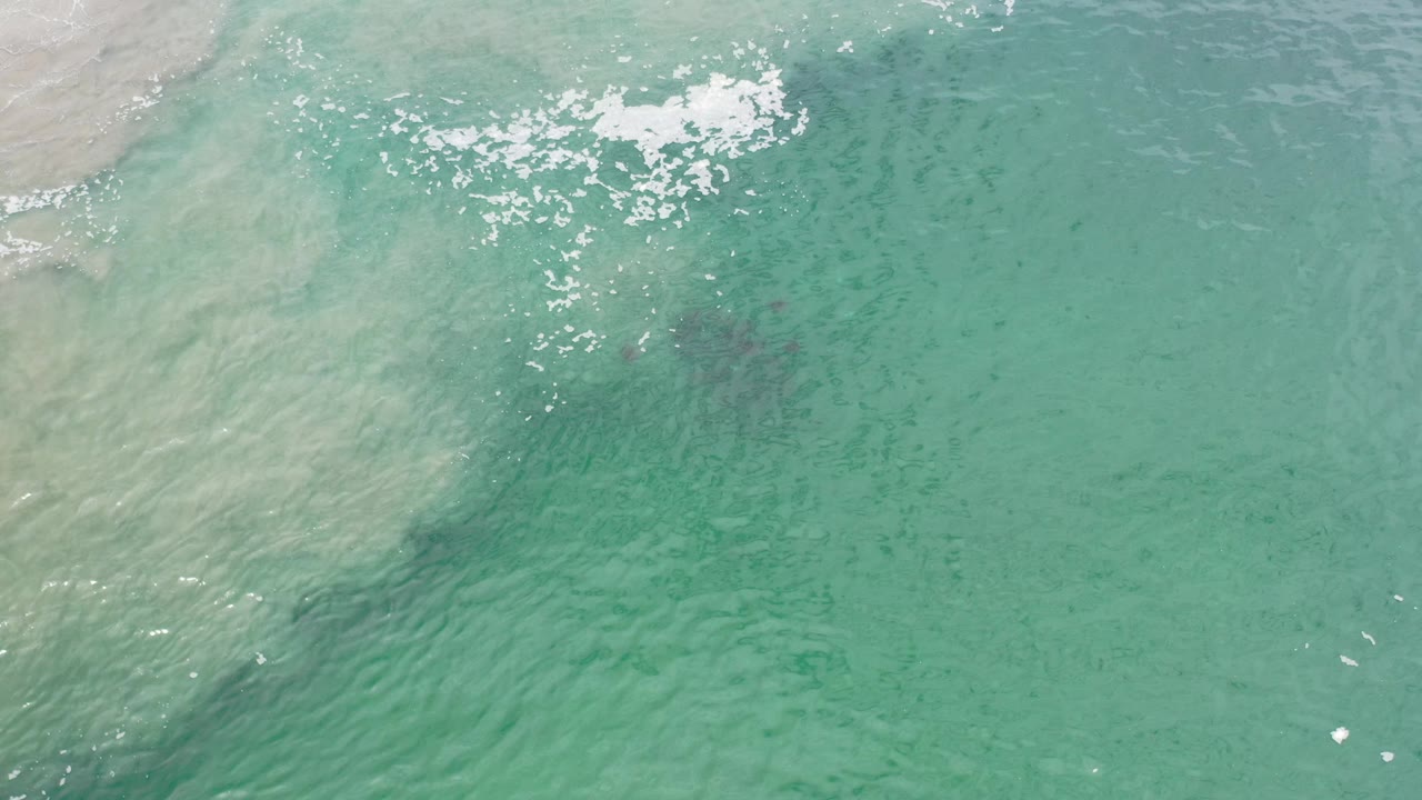 Overhead turquoise water with pale shoreline wash at upper left and white foam near the top.