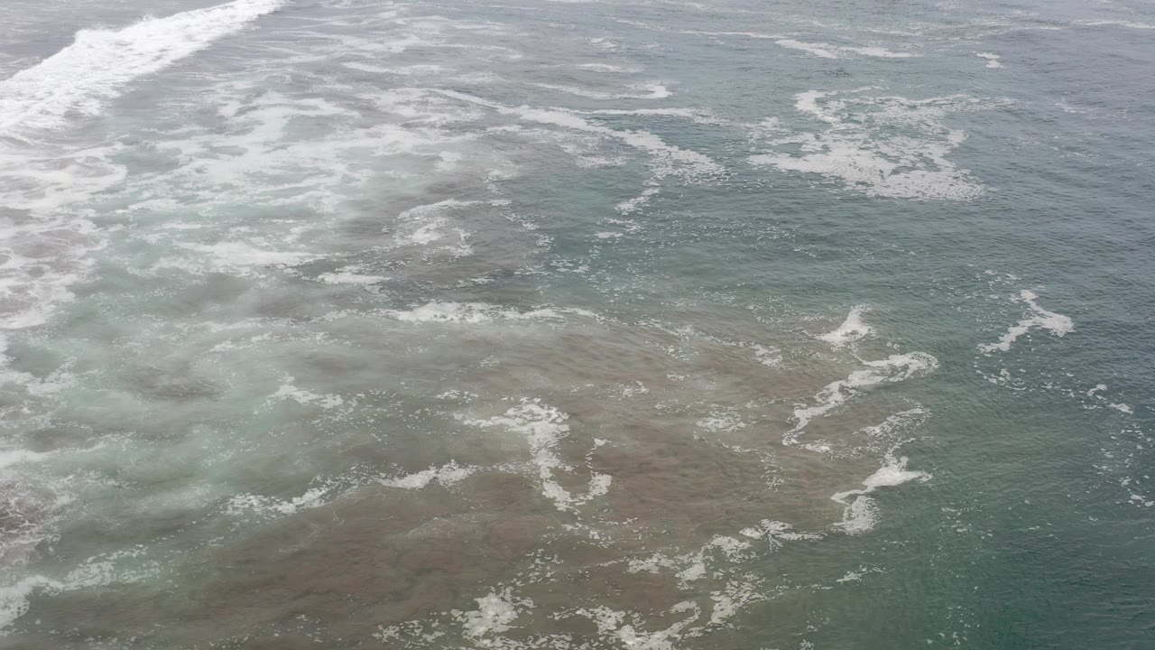 Aerial view over translucent green sea water in Los Cabos, Mexico, with white foam streaks over shallow brown reef texture and no visible people.