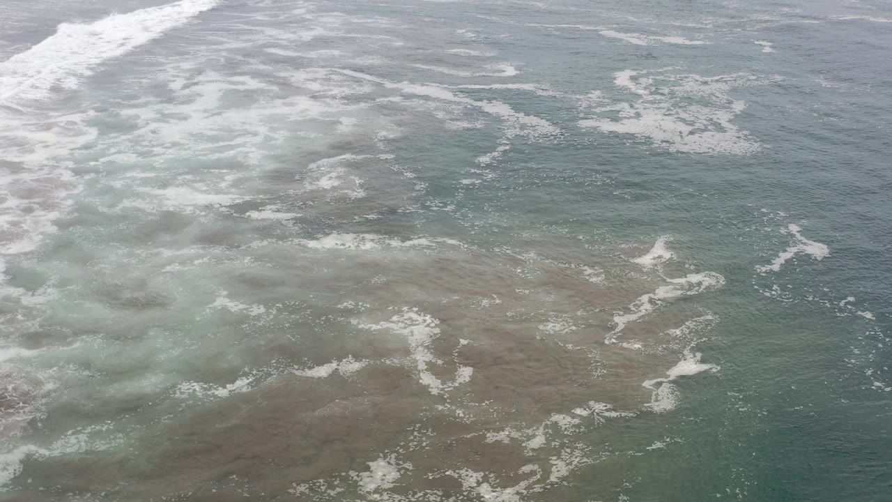 Overhead view of Los Cabos sea water where pale foam bands cross green-blue shallows above darker submerged patches, with no visible people.