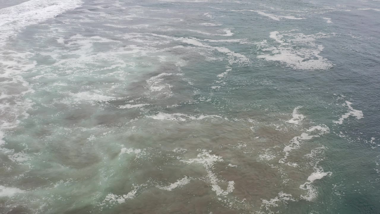 Aerial view in Los Cabos, Mexico, showing foamy white wave patterns over green-blue water and darker submerged patches, with no visible people.
