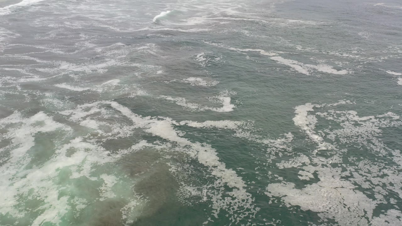 Aerial view over green-blue Los Cabos ocean water with scattered white foam and no visible people.