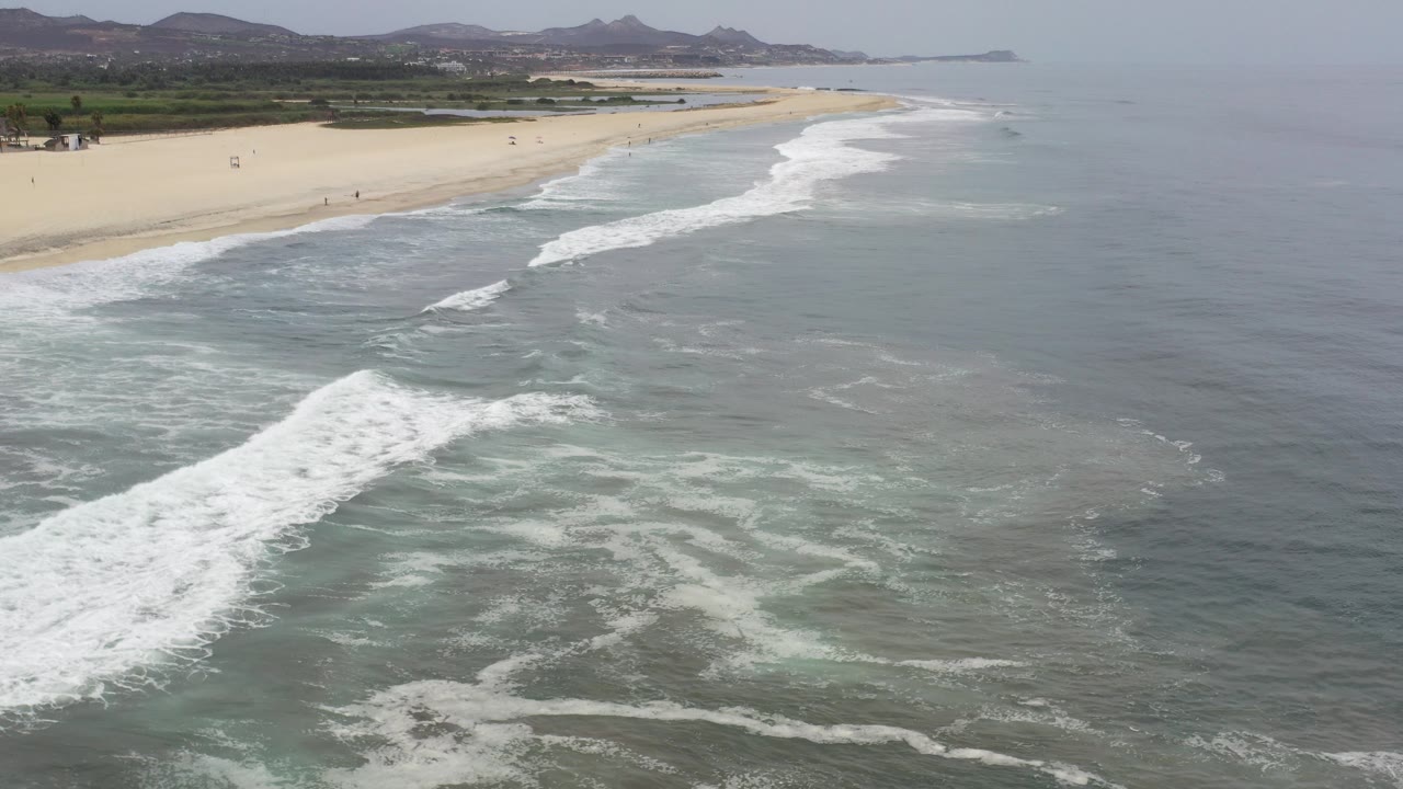 Aerial view in Los Cabos, Mexico, with white surf crossing green-blue water toward a pale sand beach, tiny distant people, low trees, and hazy mountains in the background.