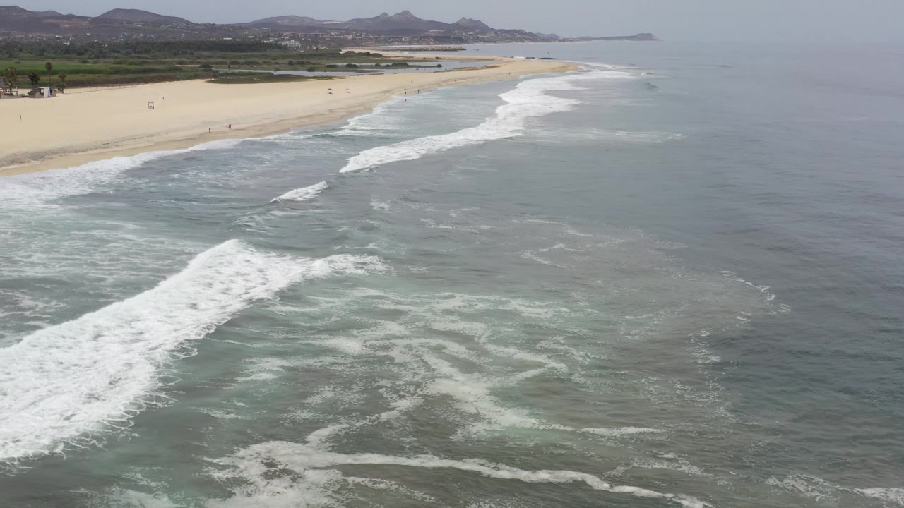 Aerial Los Cabos coastline view with a broad white wave band in the foreground, pale beach along the left, small distant beachgoers, green vegetation, and low mountains under haze.
