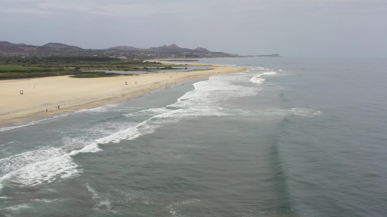 Aerial view of sandy beach, surf, and distant hills.