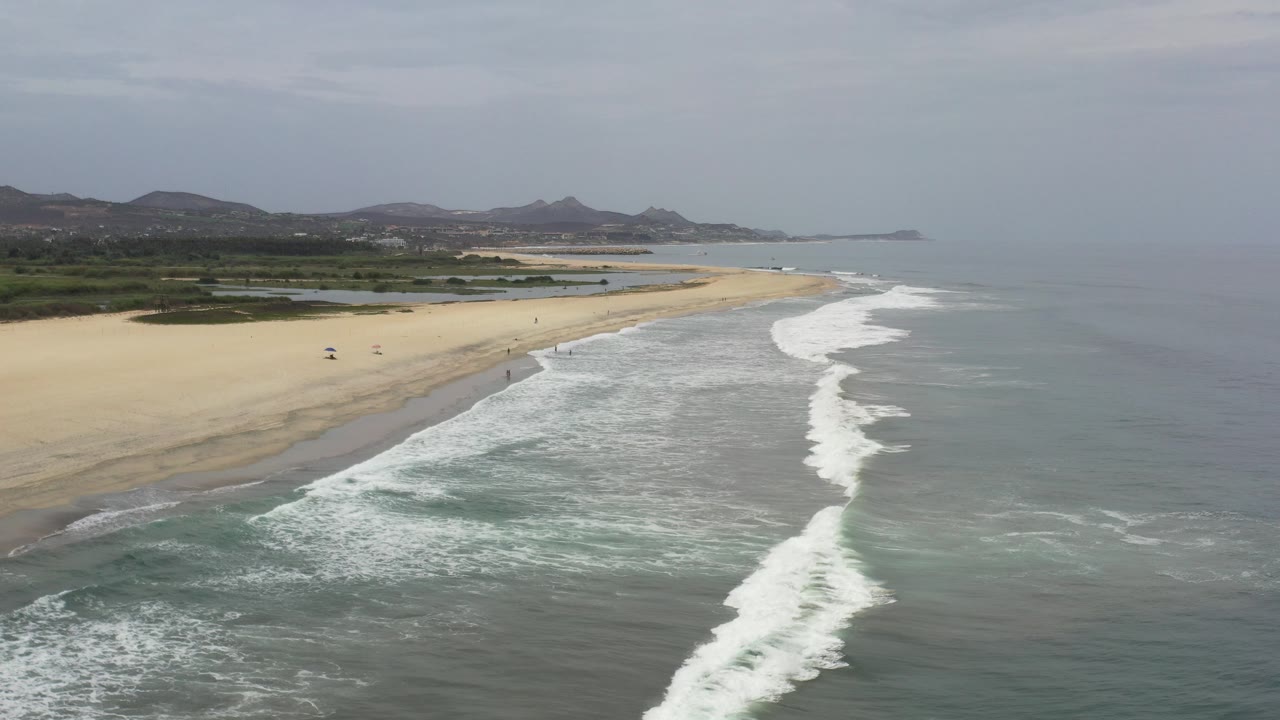 Aerial view of waves breaking along a wide beach under cloudy sky.