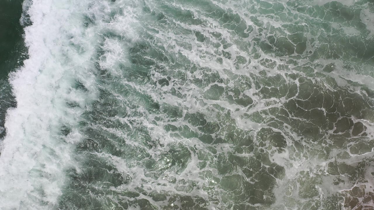 Overhead view of Los Cabos seawater with white foam crossing green gray waves at the start of the clip, with no visible people.