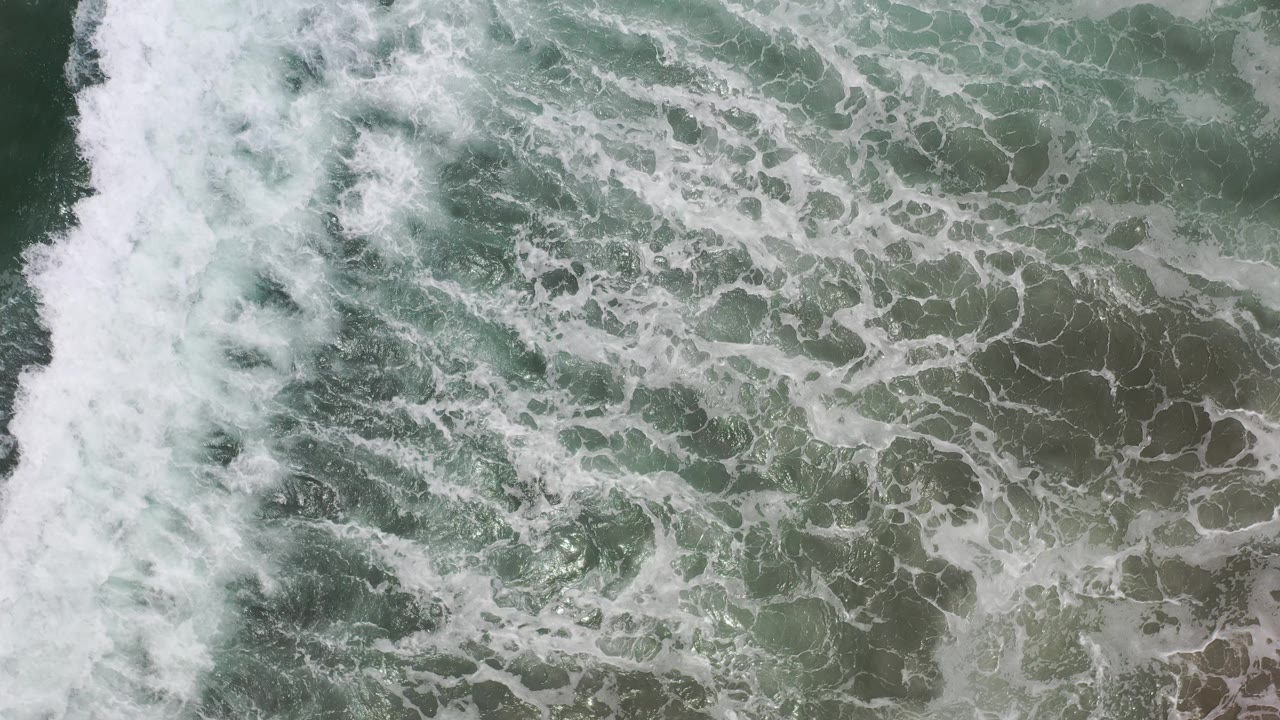 Los Cabos surf seen from above with a bright vertical band of foam against darker rippled water, with no visible people.
