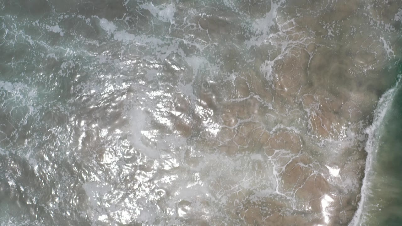 Top-down view of white foam spreading across shallow green-blue ocean water in Los Cabos, Mexico, with no visible people.