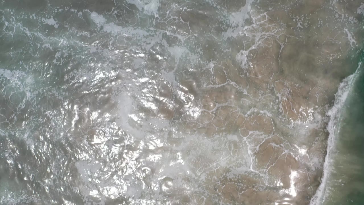 A white wave edge cuts through shallow green water in Los Cabos, Mexico, while the sandy bottom and reflected light remain visible, with no visible people.