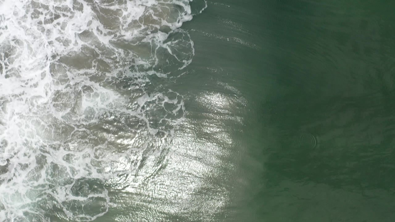 Overhead view of Los Cabos ocean water with white surf foam entering from the upper left and a bright sun reflection on green ripples, with no visible people.