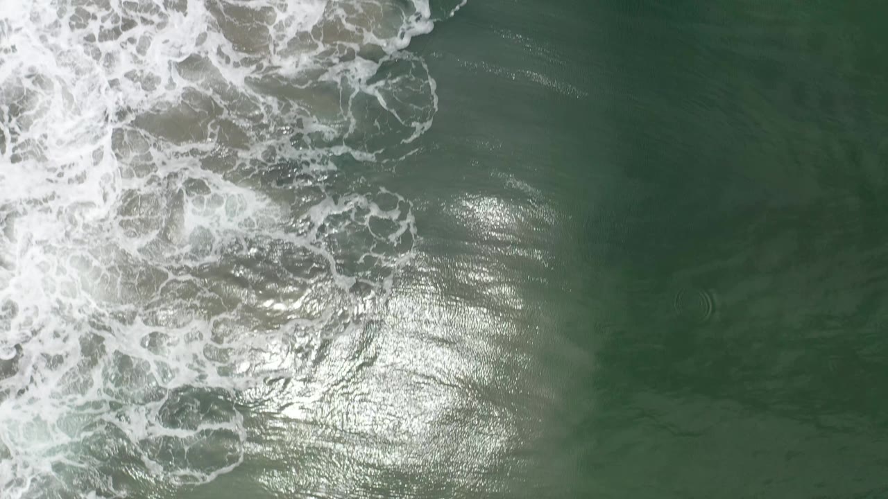 Top-down Los Cabos surf frame with a thicker band of white wave foam crossing the upper left and sunlight shimmering across darker green water, with no visible people.