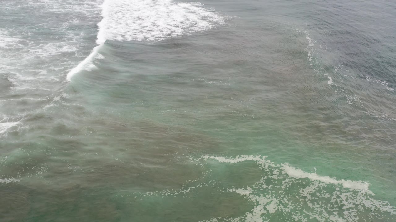 High oblique view of white surf breaking across teal-gray ocean water in Los Cabos, Mexico, with foamy water in the foreground and no visible people.