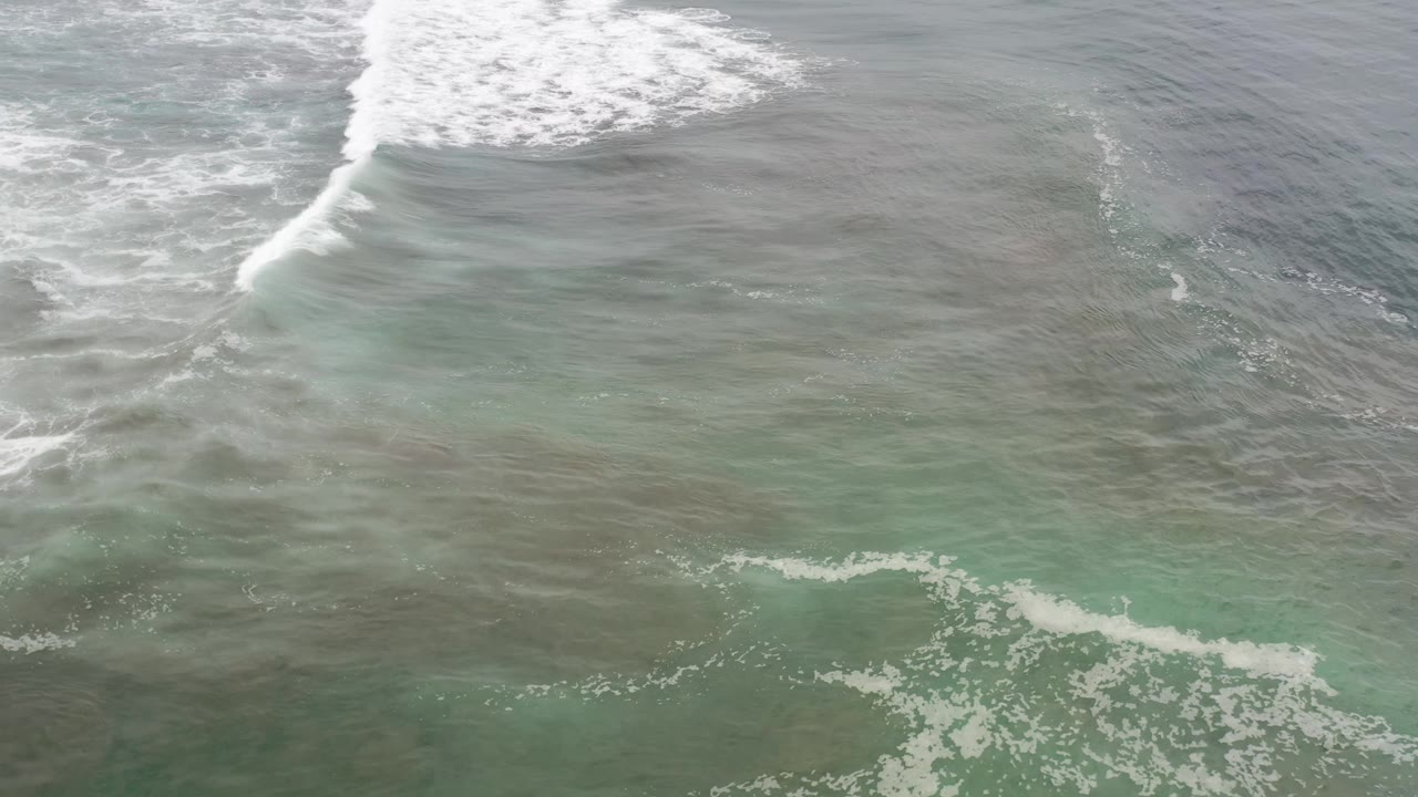 A diagonal band of white surf crosses shallow green-gray water in Los Cabos, Mexico, with the beach outside the frame and no visible people.