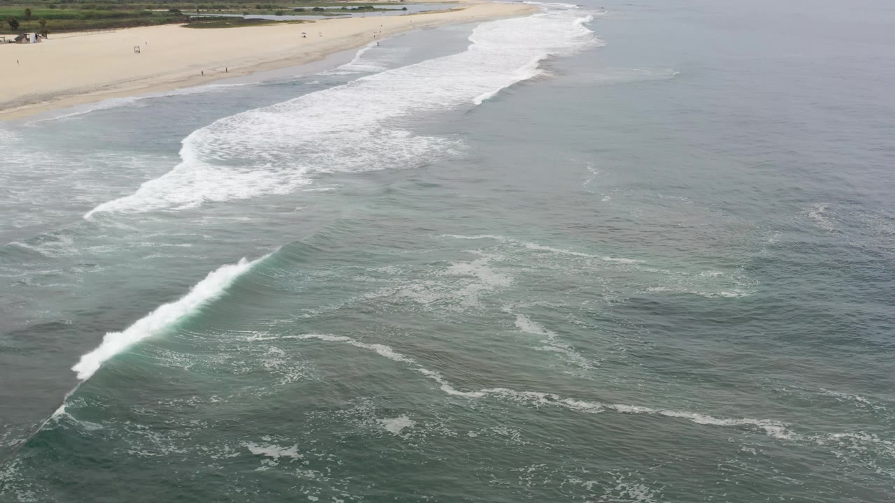 High aerial frame of Los Cabos surf forming a diagonal white break beside a wide sandy beach and muted ocean, no visible people.