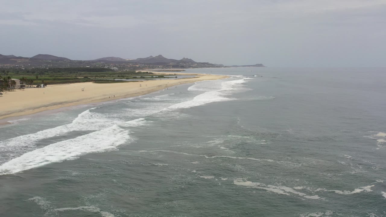 Aerial view of gray ocean waves reaching a sandy beach with hills beyond.