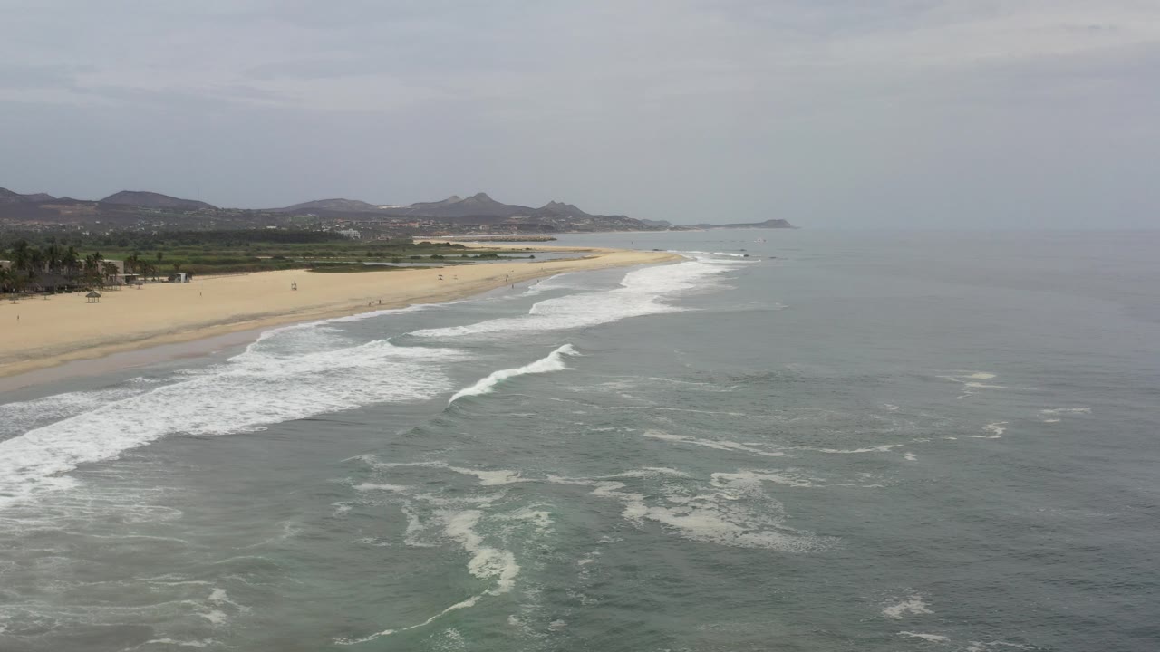Wide aerial view of a Los Cabos, Mexico beach with gray-green ocean in the foreground, white surf bands along the sand, distant low mountains, and no clearly visible people.