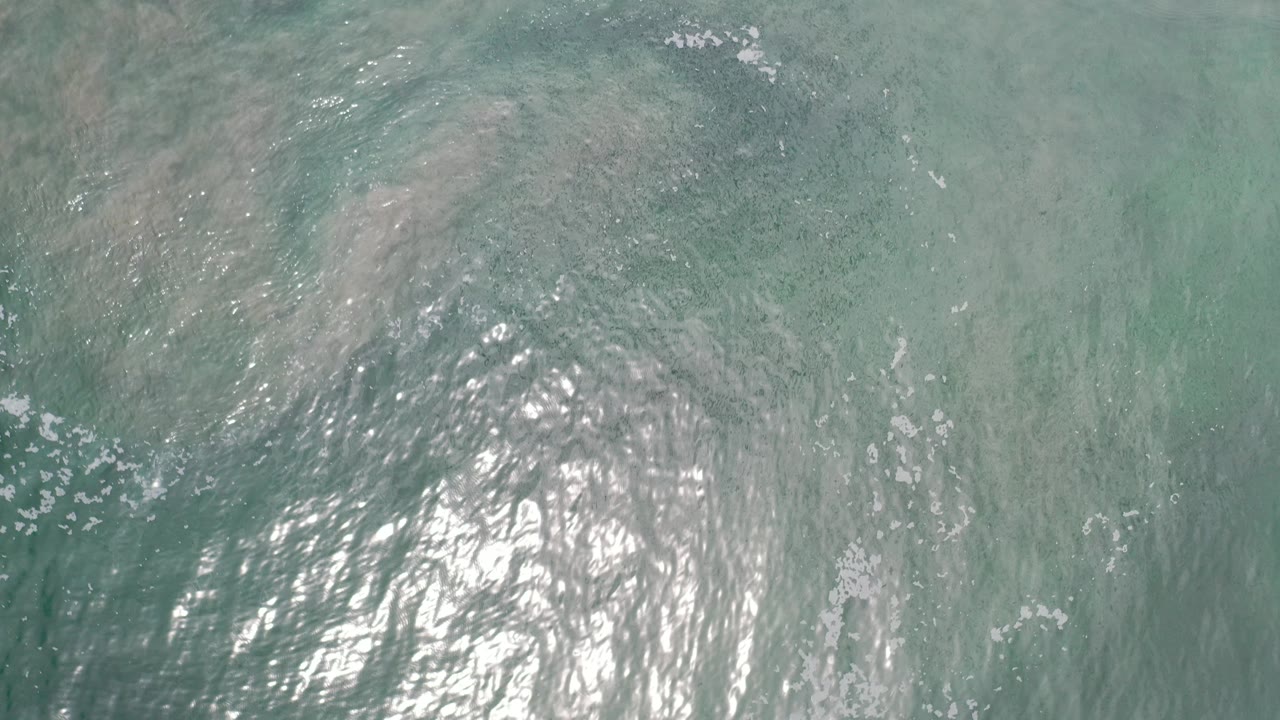 Overhead view of pale turquoise ocean in Los Cabos, Mexico, with bright sun glare and darker submerged patches, no visible people.