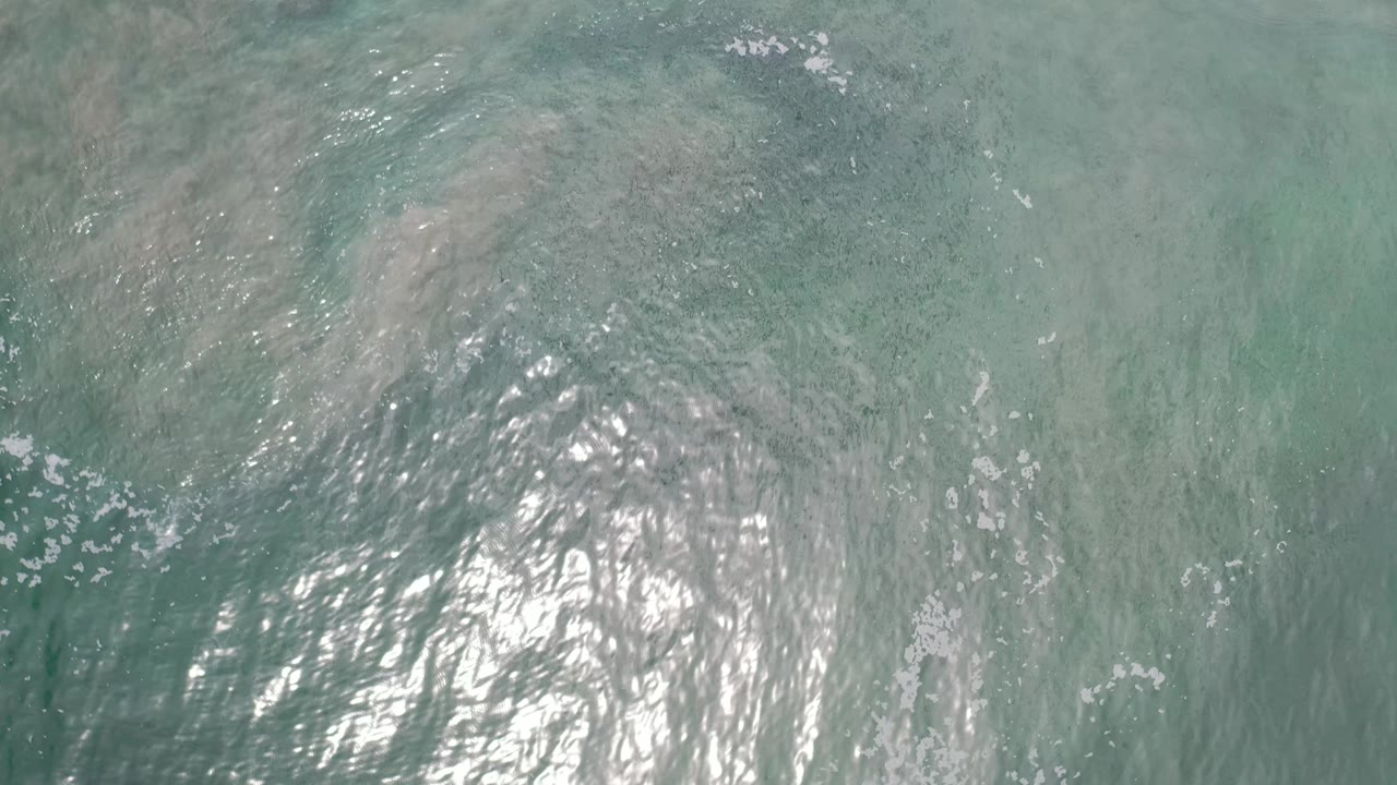 Aerial view over Los Cabos ocean water with a concentrated patch of sun reflection near the lower center and faint darker bands below the surface, no visible people.