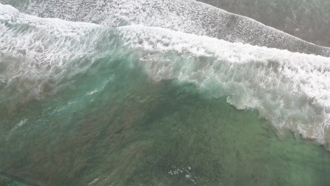 Overhead view in Los Cabos, Mexico of white surf spreading across green-blue water and tan shoreline, with no visible people.
