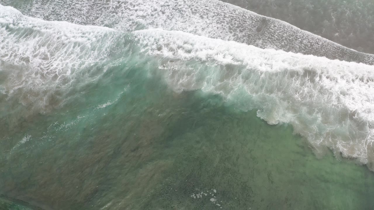 Aerial frame in Los Cabos, Mexico with a broad whitewater wave line crossing shallow green ocean above a narrow strip of sand, with no visible people.