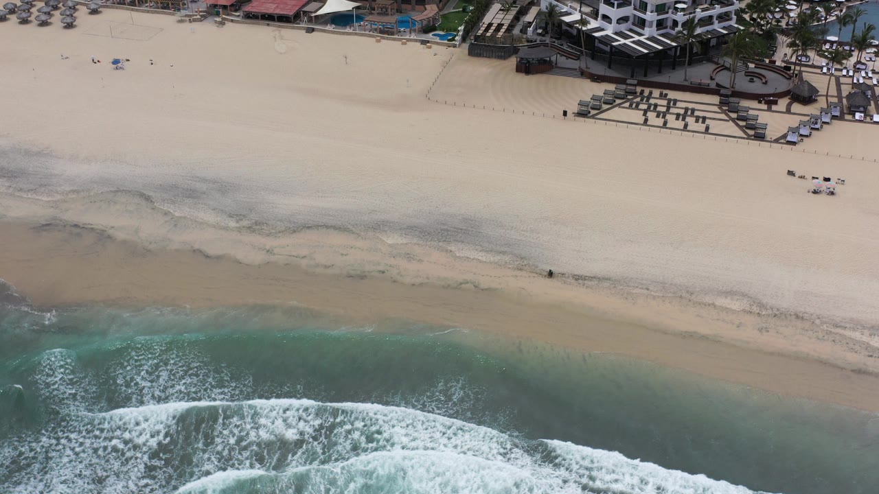 Aerial frame over Los Cabos, Mexico showing turquoise surf, pale sand, beach seating, and the edge of a beachfront hotel complex with no visible people.