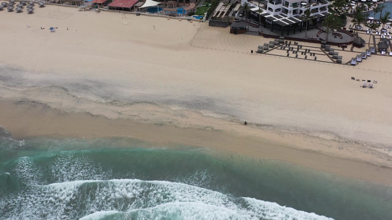 Aerial frame over Los Cabos, Mexico with a broad band of sand, breaking waves, terraced seating, and hotel roofs along the top of frame with no visible people.