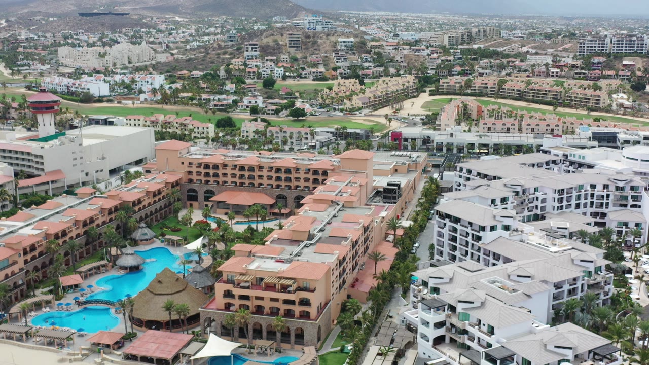 Los Cabos aerial frame with white condo rooftops at right, terracotta resort blocks and pools at left, hillside neighborhoods beyond, and no visible people.