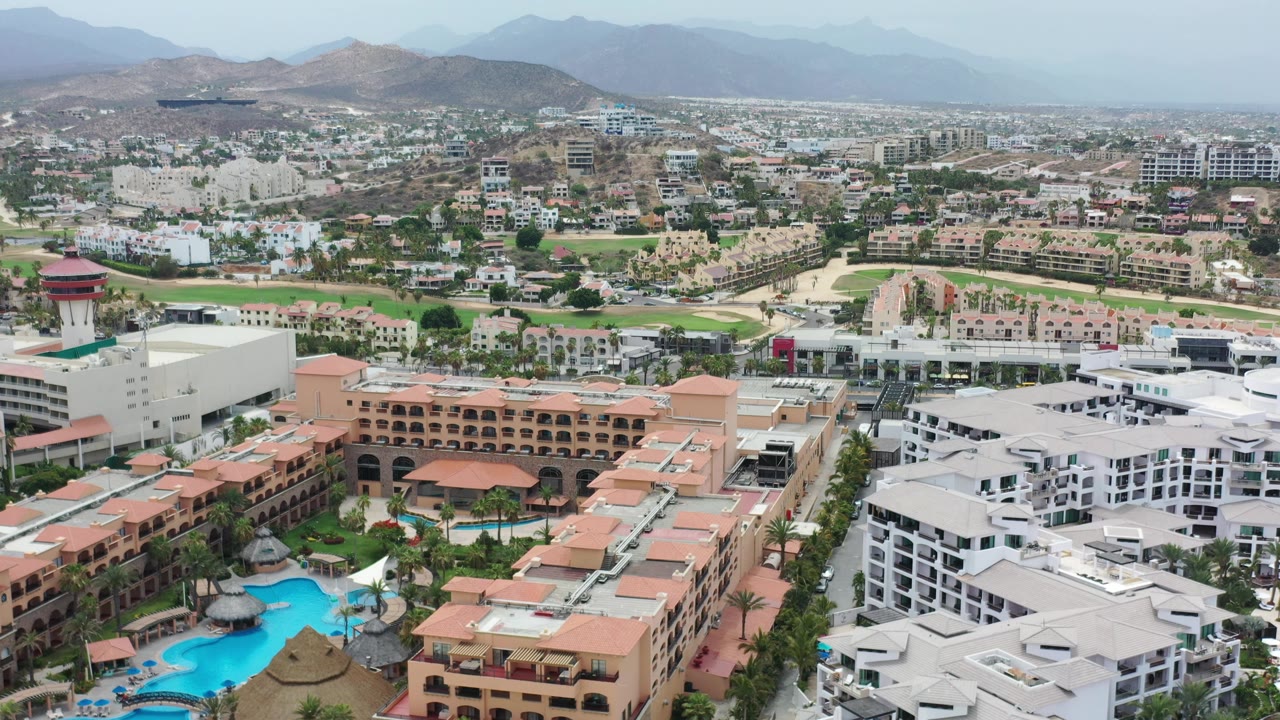 Wide Los Cabos view showing a pool complex and white residential buildings in the foreground, dense development in the valley, layered mountains, and no visible people.
