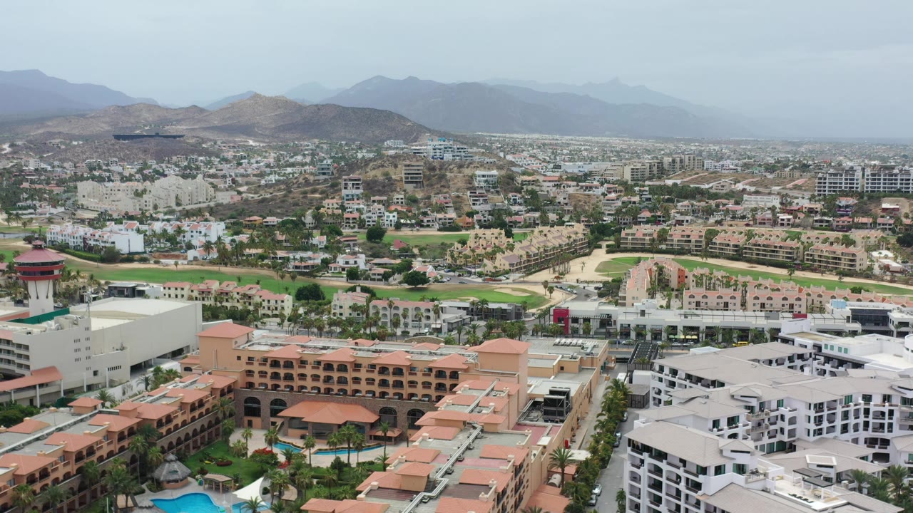 Wide aerial of resort rooftops, pools, roads, green fairways, and distant hills.