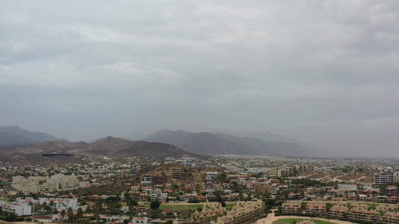 Los Cabos city blocks and green patches sit below a continuous gray sky, with dark hills across the middle distance, hazy mountains behind them, and no visible people.