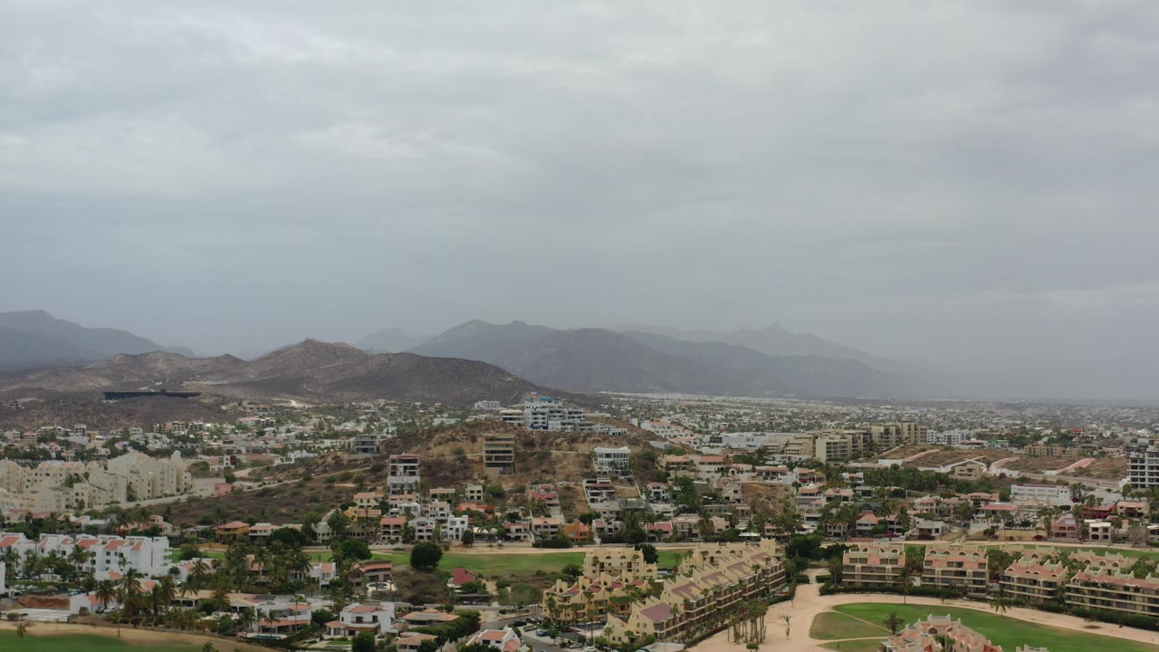 High aerial view over buildings, roads, green grounds, dry hills, and cloudy sky.