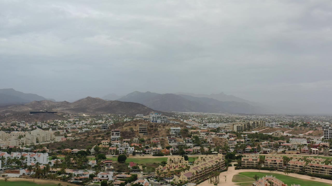 Los Cabos aerial view looking over clustered homes and larger buildings toward dry hills and a hazy mountain ridge, with no visible people in the frame.
