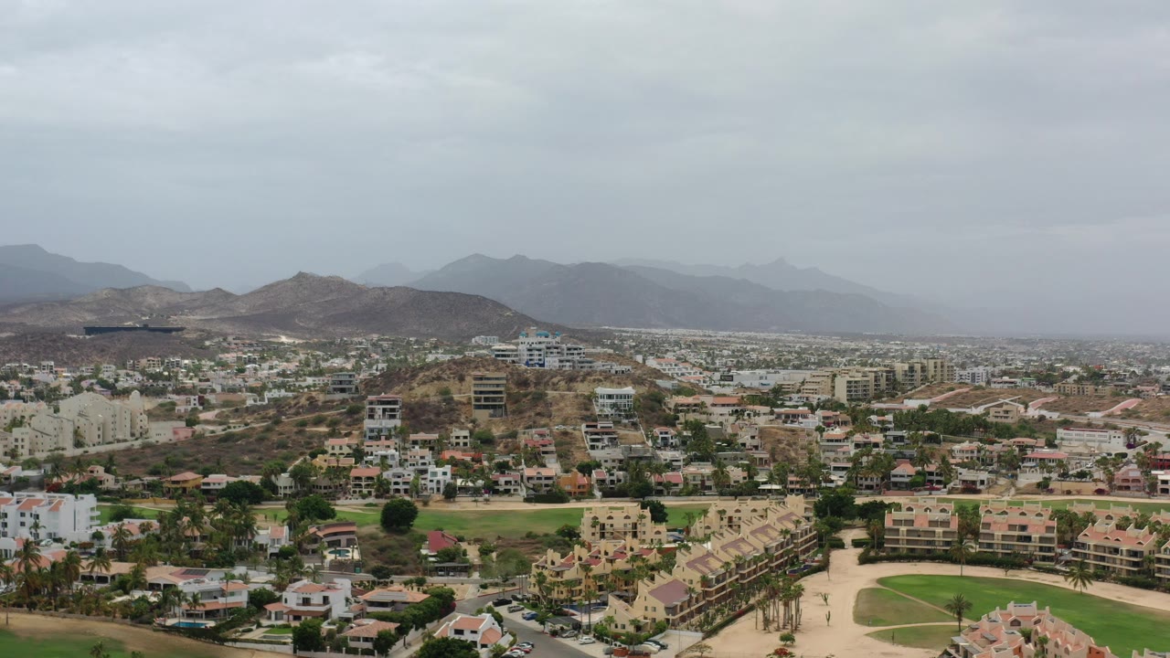 Wide aerial view of Los Cabos buildings, rooftops, hills, and cloudy sky.
