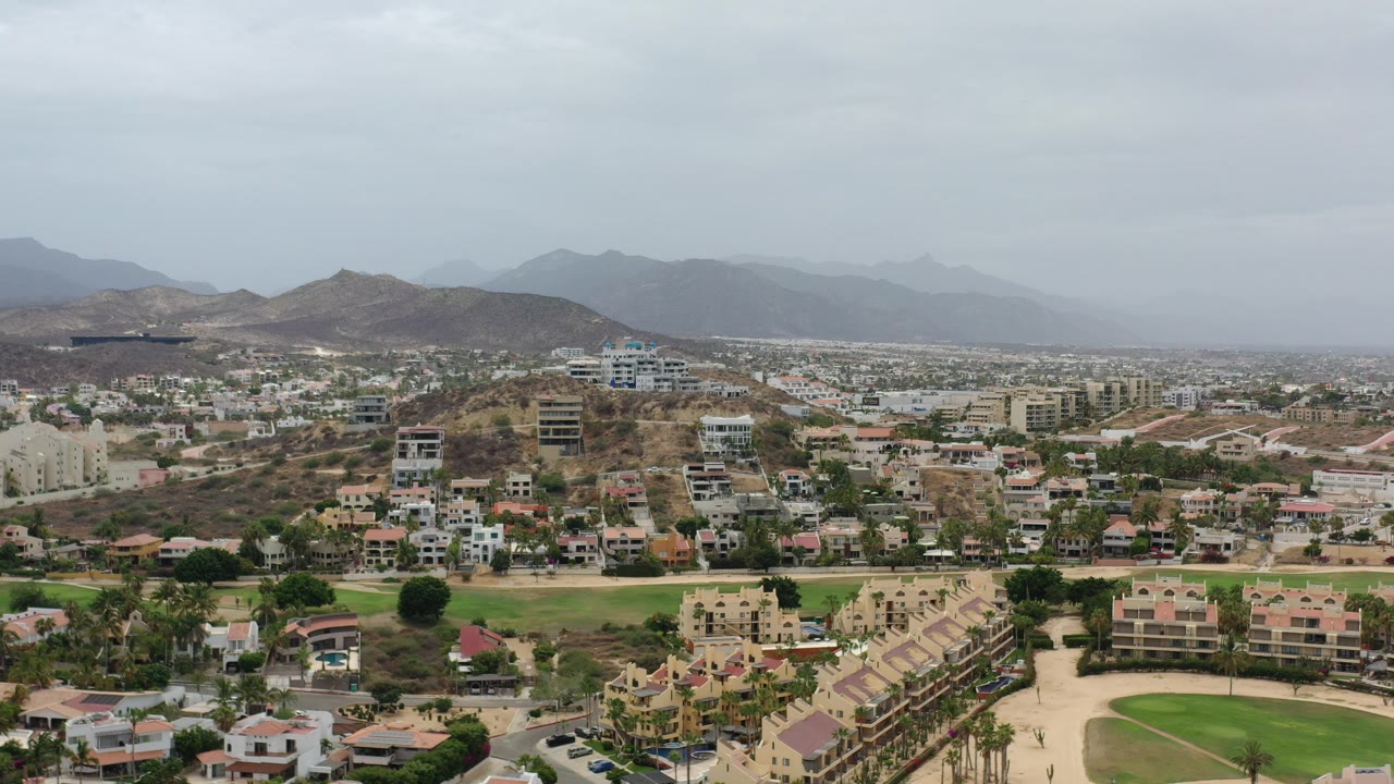Opening aerial frame of Los Cabos, Mexico, showing hillside homes, roads, palm landscaping, arid slopes, distant mountains, and no visible people.
