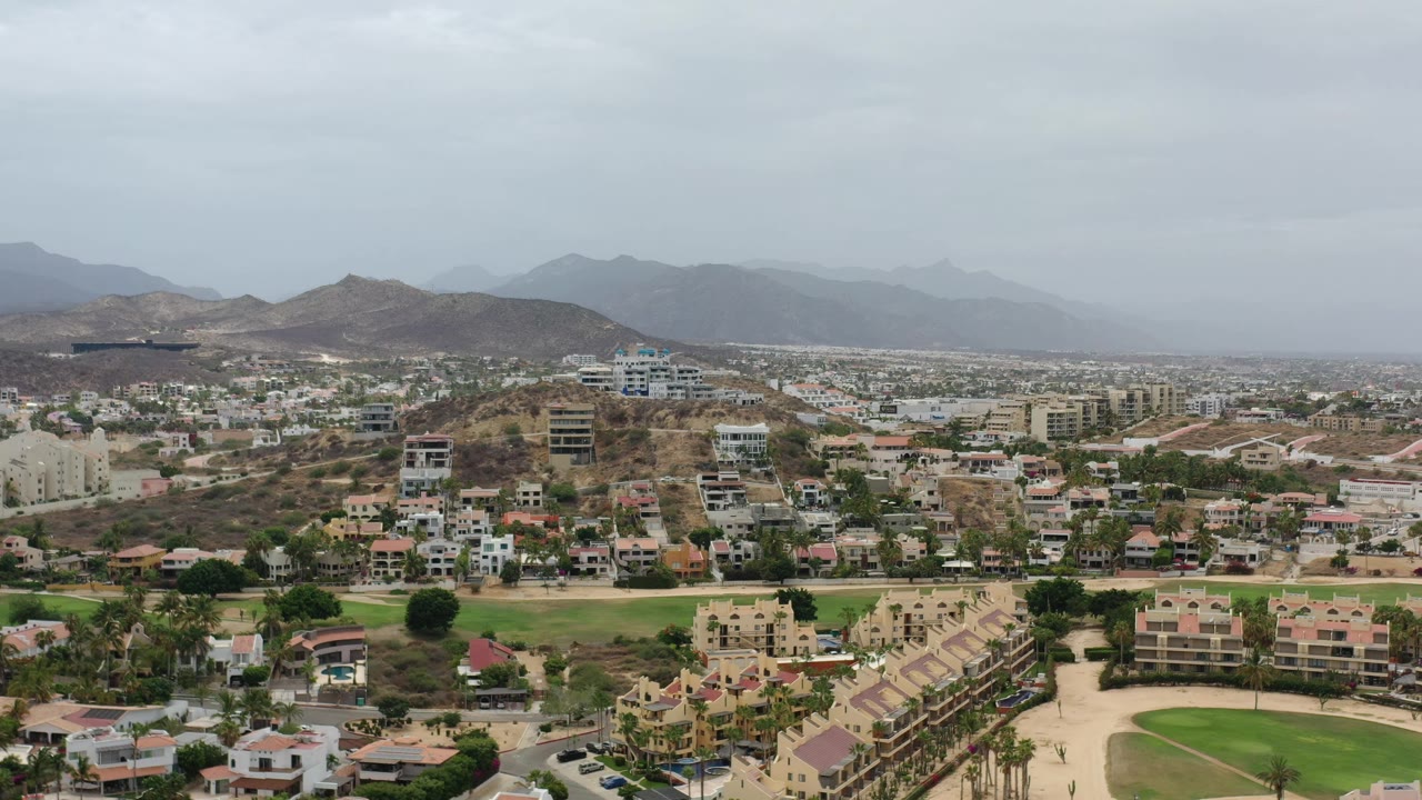 Aerial Los Cabos, Mexico view with residential blocks, a broad green landscaped area, nearby roads, brown hills, hazy mountains, and no visible people.