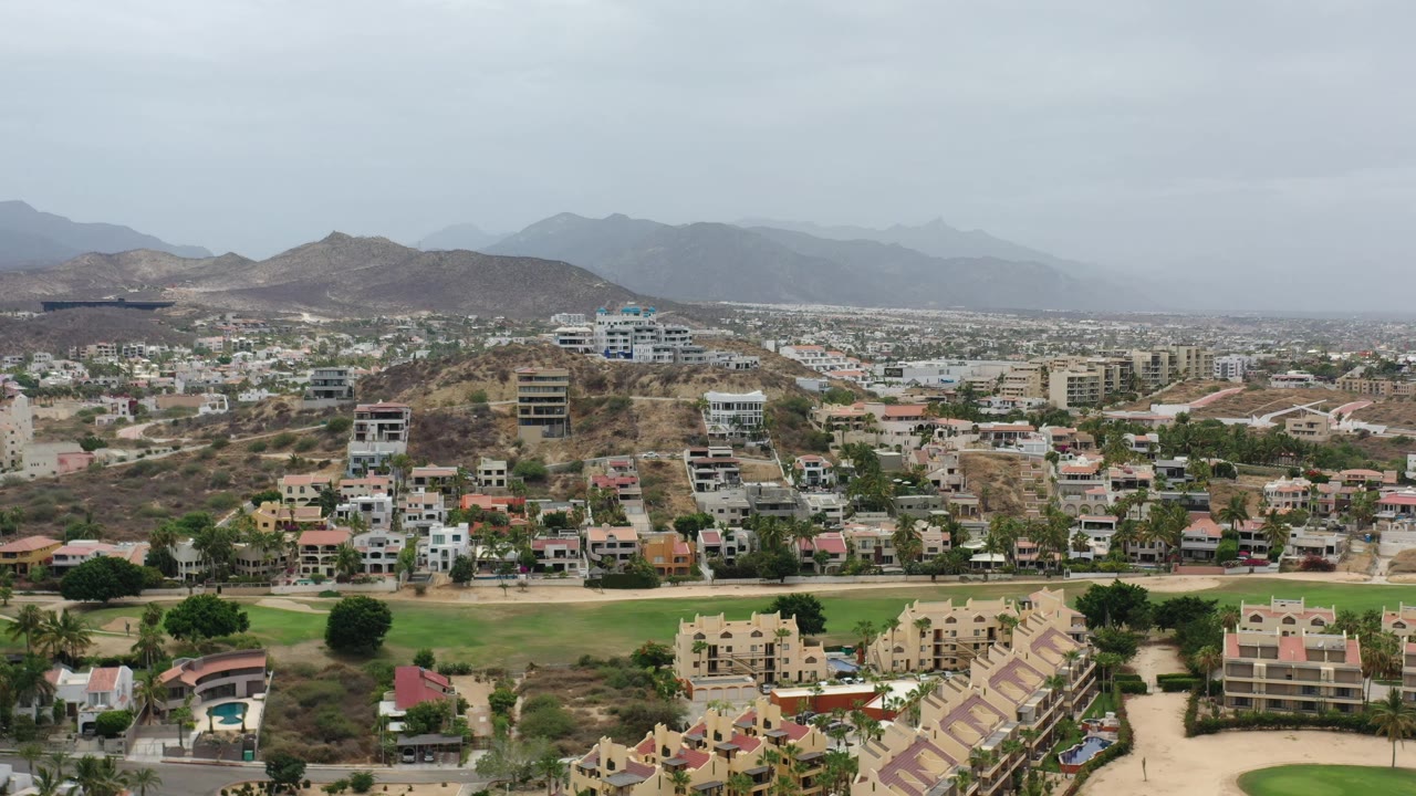 Aerial view in Los Cabos, Mexico with residential buildings spread across low hills, a green course-like strip below, hazy mountain ridges, and no visible people.