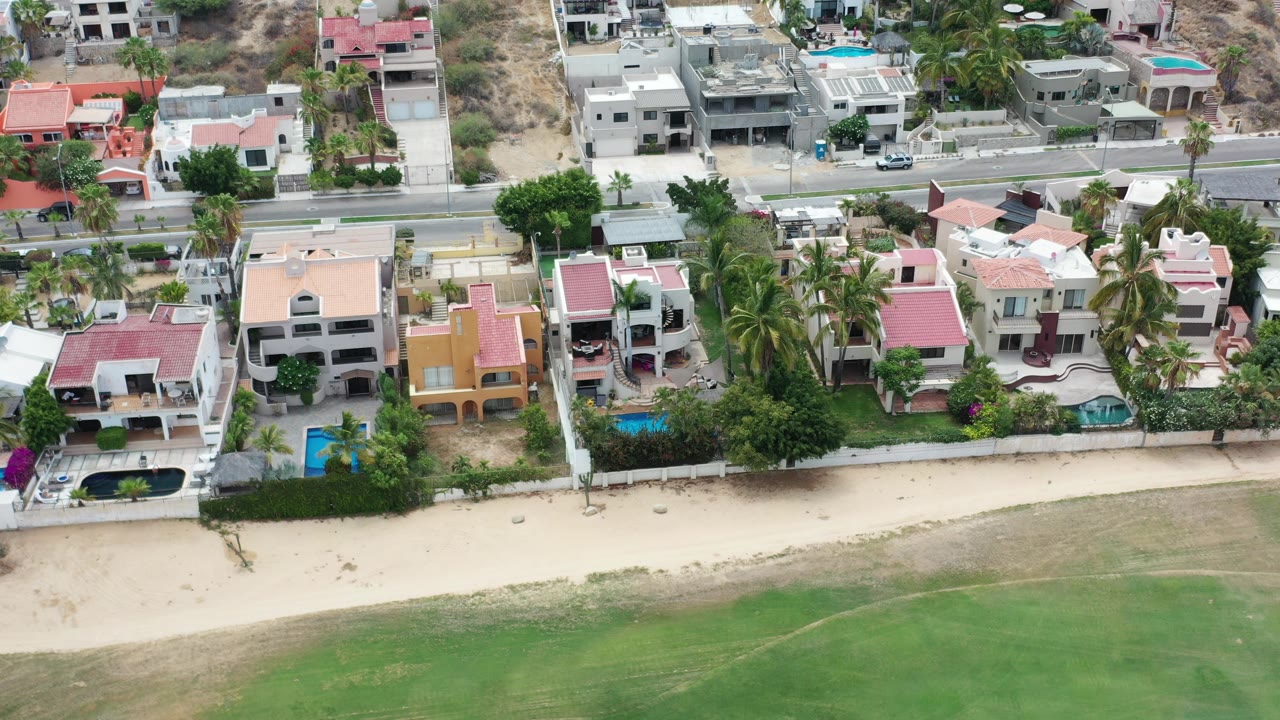 Aerial view of beachfront houses, pools, sand, green water, and road.