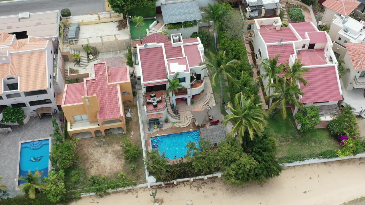 Second overhead frame in Los Cabos, Mexico with the camera view shifted across adjoining homes, one bright turquoise pool, one darker blue pool, palms, patios, and no visible people.