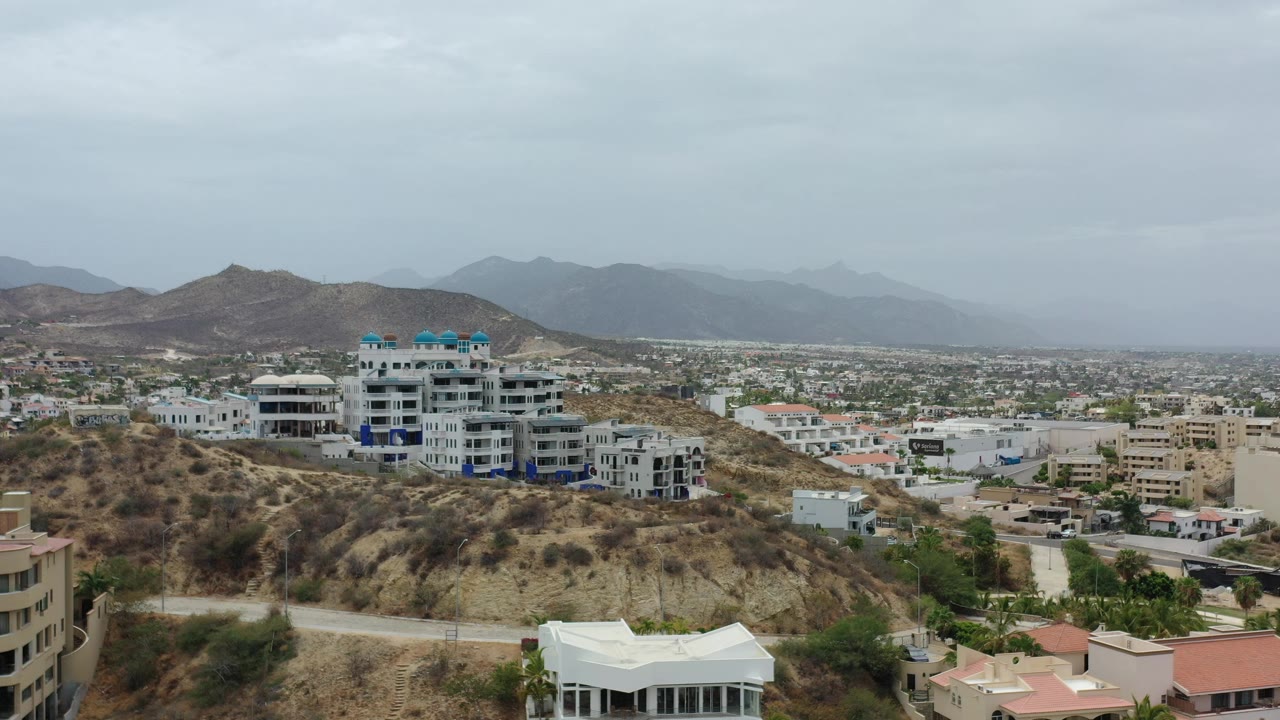 Opening frame of Los Cabos, Mexico with hillside buildings, dry scrub terrain, hazy mountain ridges, gray sky, and no visible people.