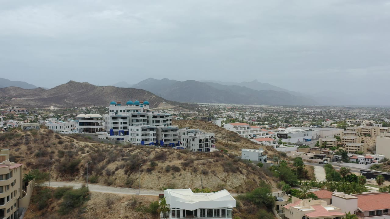 Los Cabos hillside view with a prominent white multi-story building, beige city structures, dry slopes, distant mountains, and no visible people.