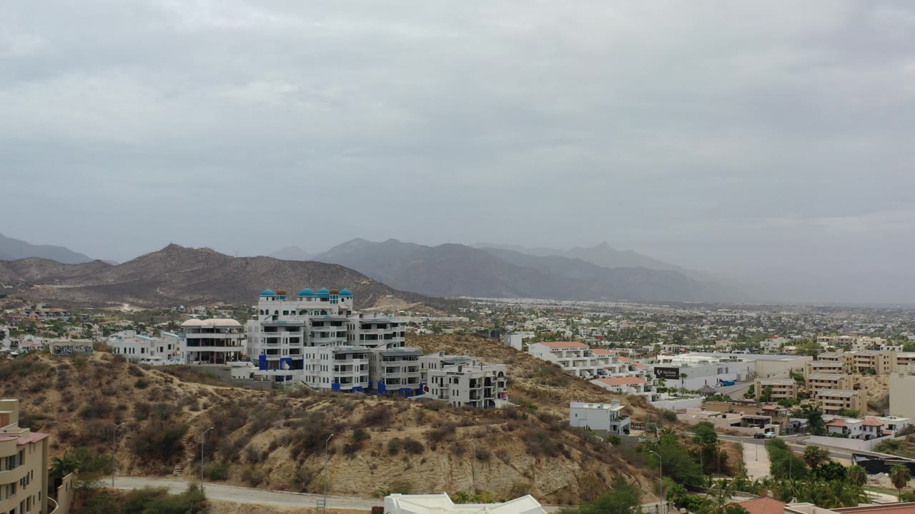 Wide hillside view with white condos, houses, city buildings, mountains, and cloudy sky.
